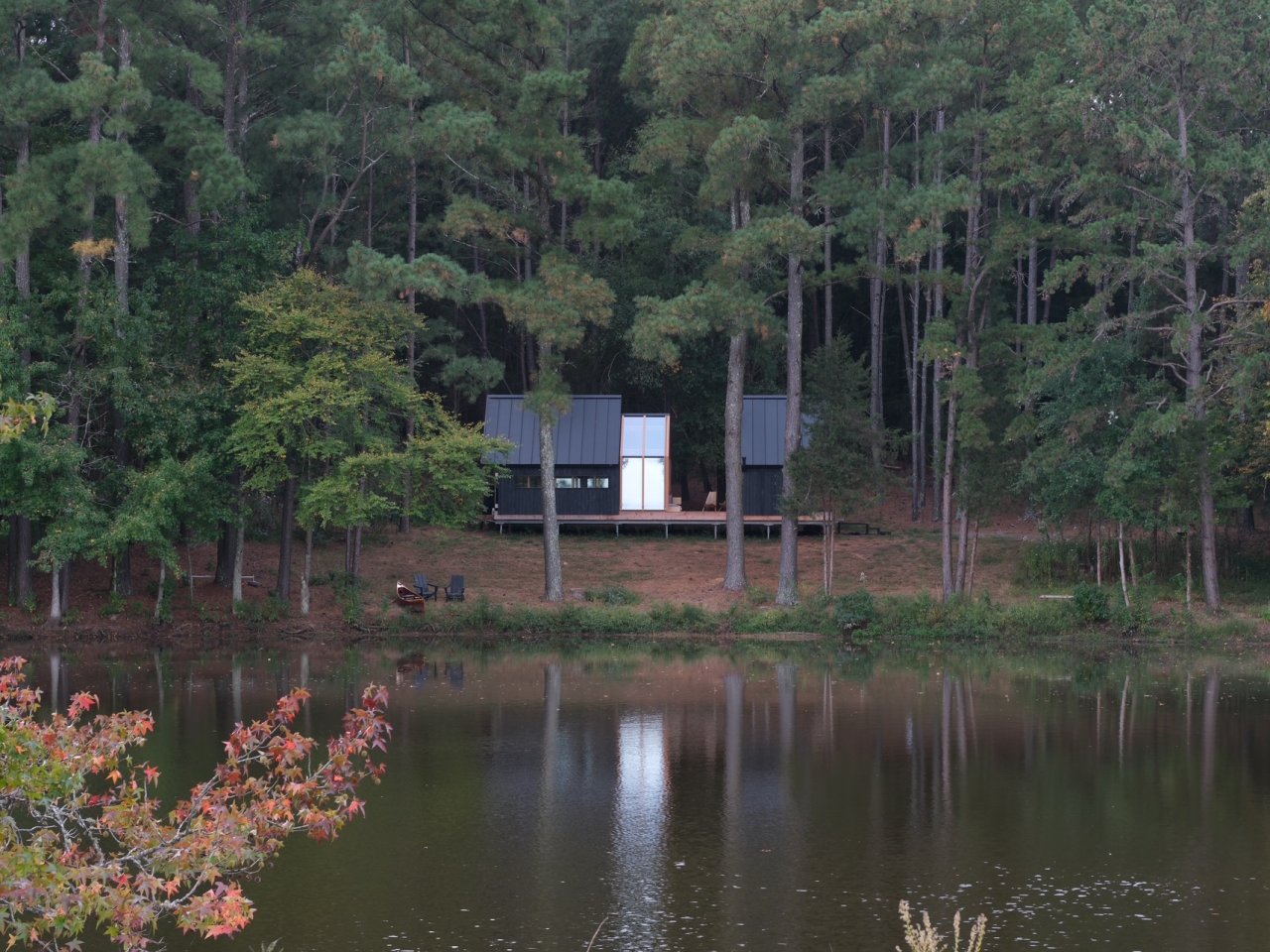 Cabin ANNA set among tall pine trees beside a quiet pond, with its dark timber exterior and central glass shell reflected in the water.