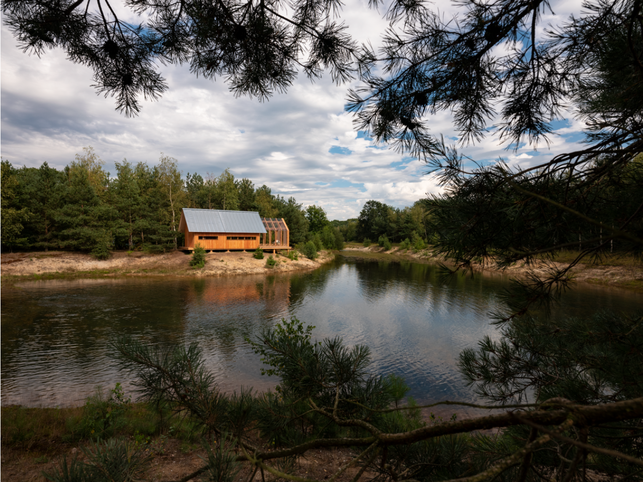 Cabin ANNA set beside a secluded pond, surrounded by pine forest and framed by tree branches.