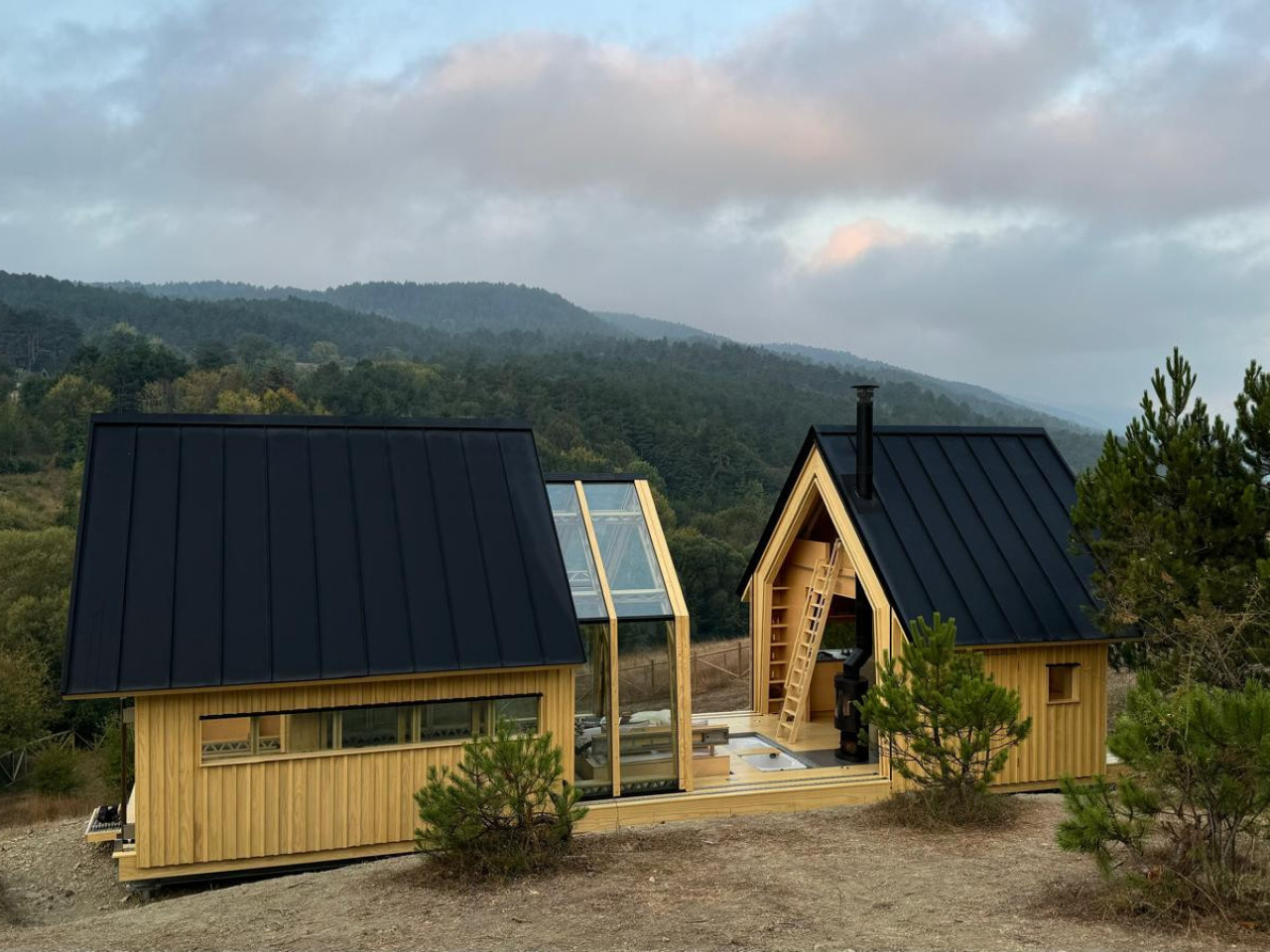Cabin ANNA opened to the surrounding mountain landscape, with its timber shells and central glass structure overlooking forested hills.